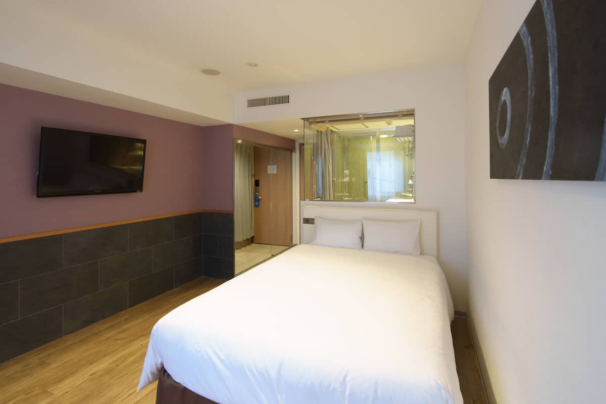 A hotel room with a large bed, a flat-screen TV mounted on the wall, a large mirror above the bed, and a wooden floor.