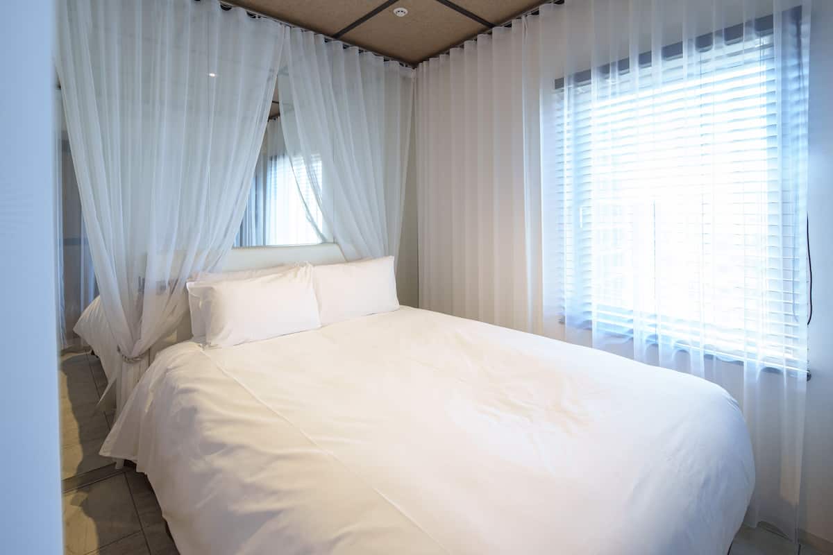 A large bed with white linens, sheer curtains, and a window with blinds.