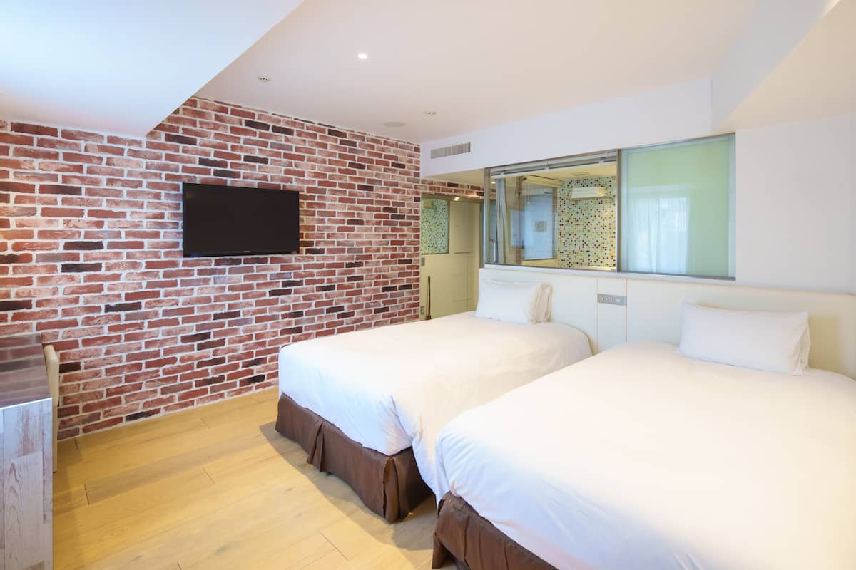 A hotel room with two beds, a brick wall, a flat-screen TV, and a large window.
