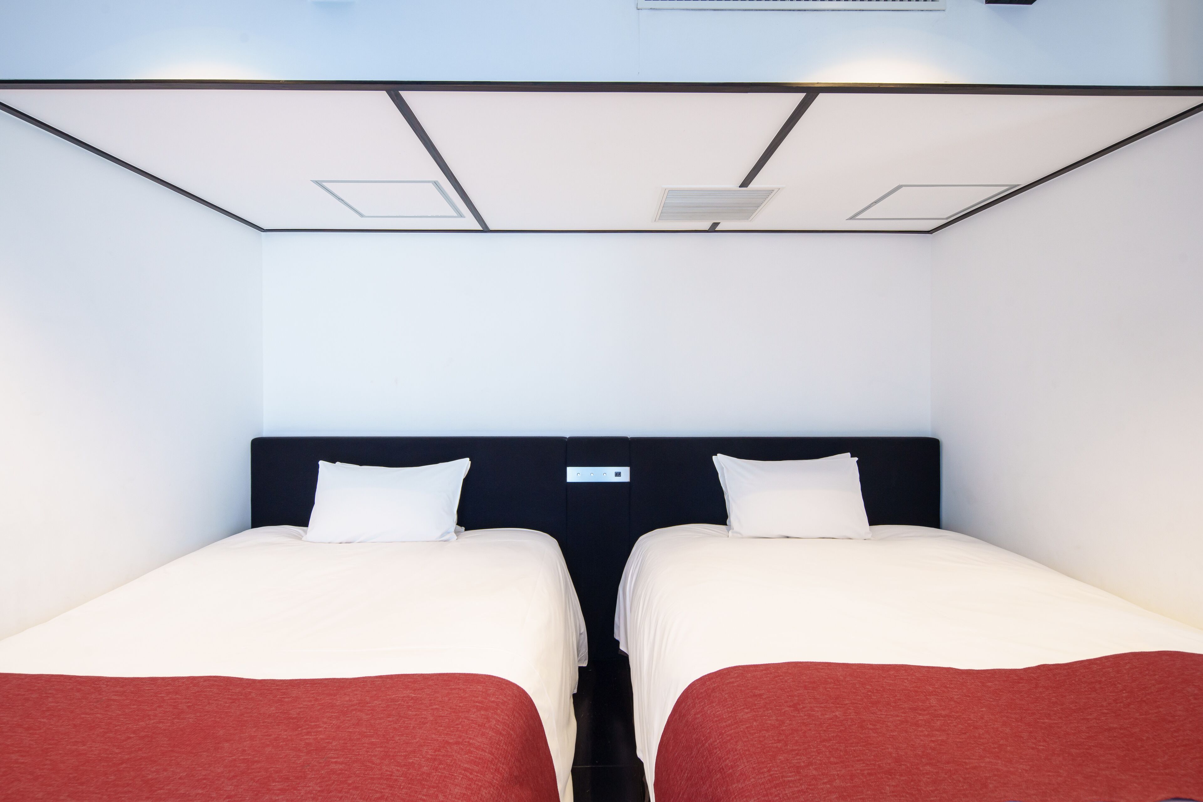Een hotelkamer met twee bedden, elk voorzien van witte lakens en rode plaids. De kamer heeft een modern design, met een zwart hoofdbord en een plafond met inbouwverlichting.