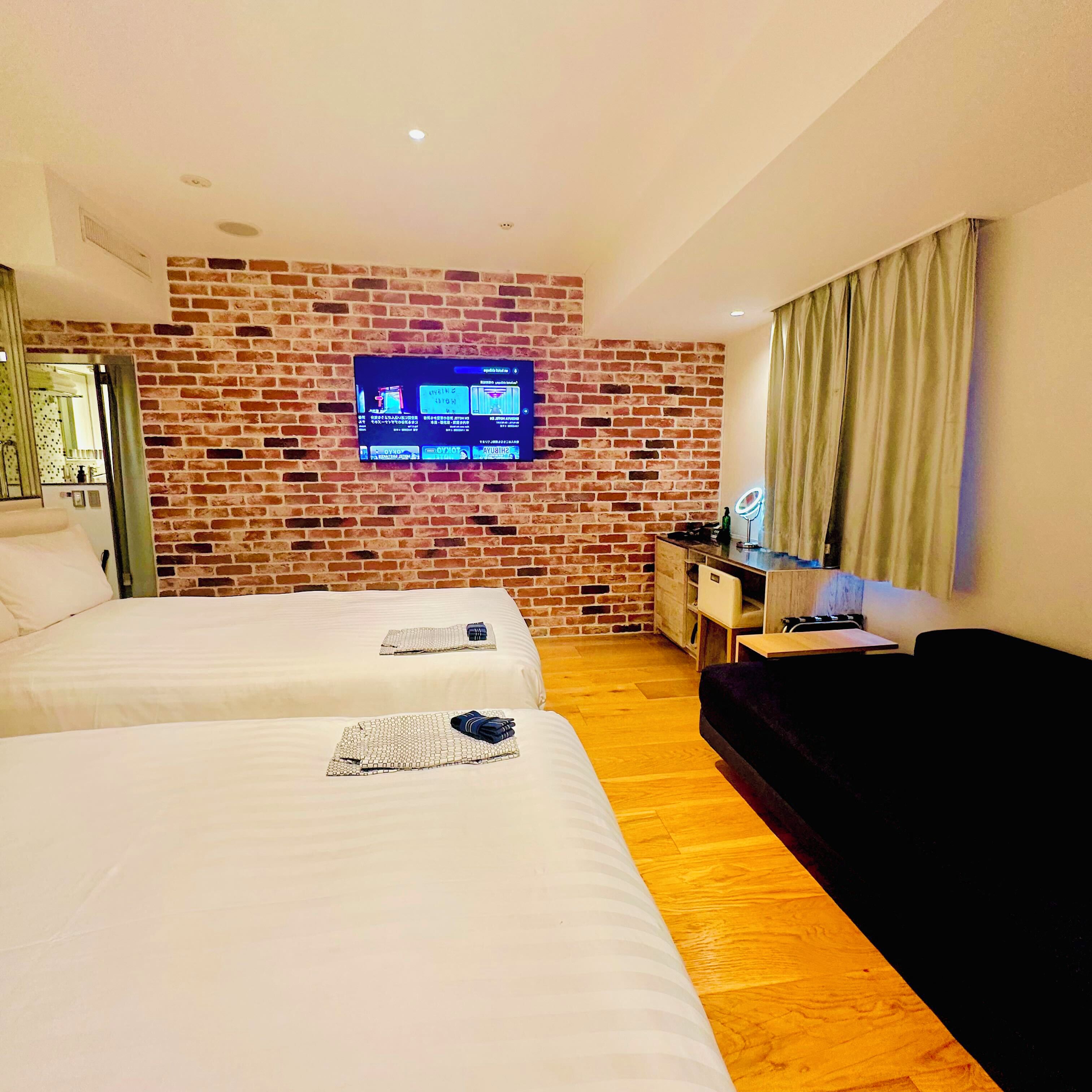 A hotel room with two beds, a brick wall, a television, a desk, and a sofa.