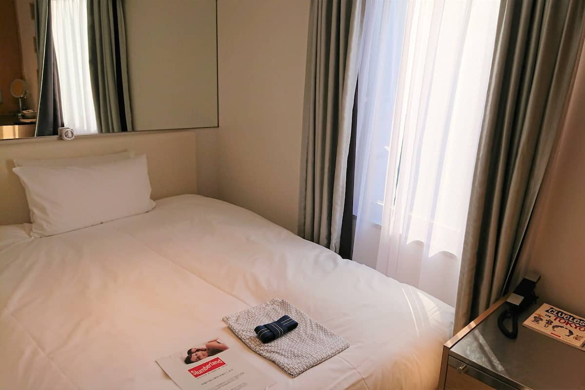 A hotel room with a neatly made bed, a bedside table with a lamp, and a window with curtains.