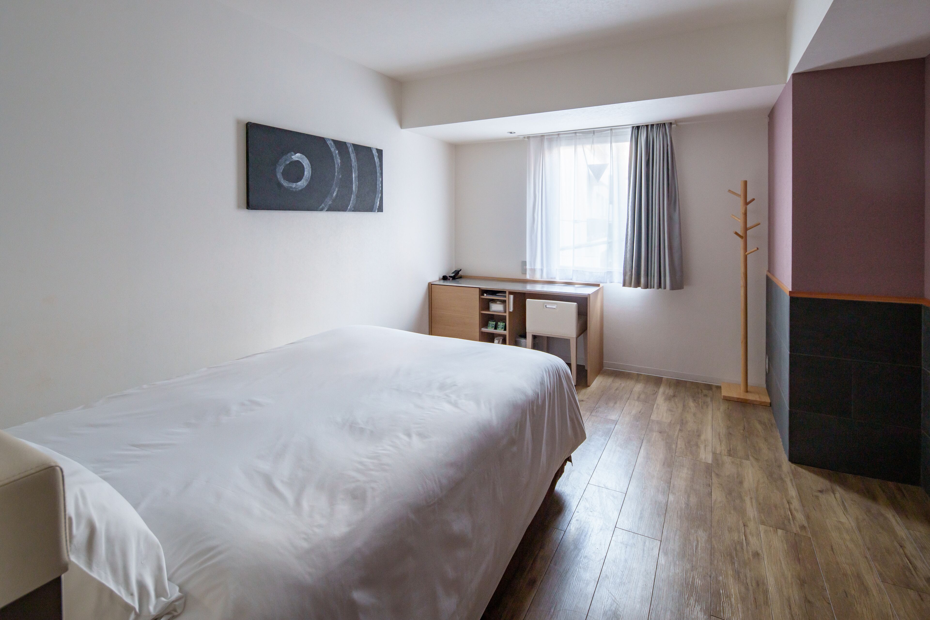 A bedroom with a bed, a desk, a coat rack, and a window with curtains.
