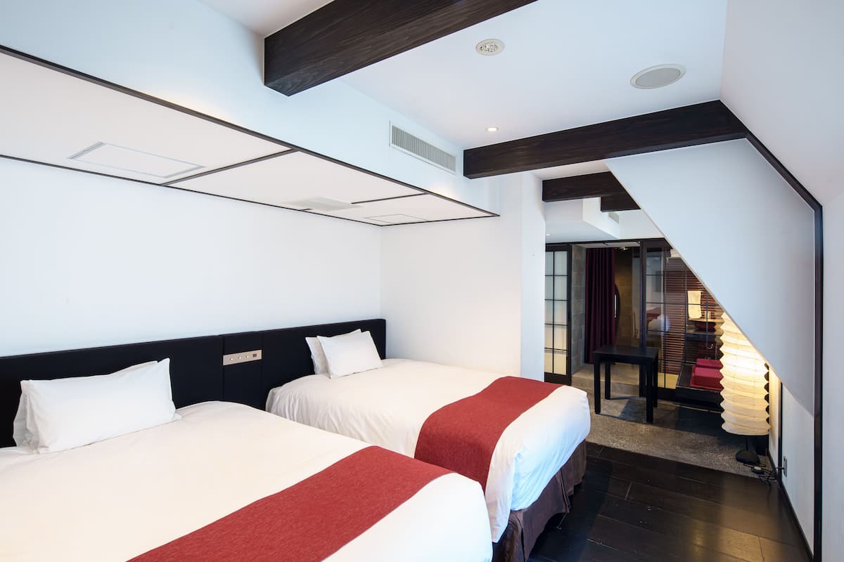 A hotel room with two beds, a dark wooden ceiling, and a staircase leading to a common area.