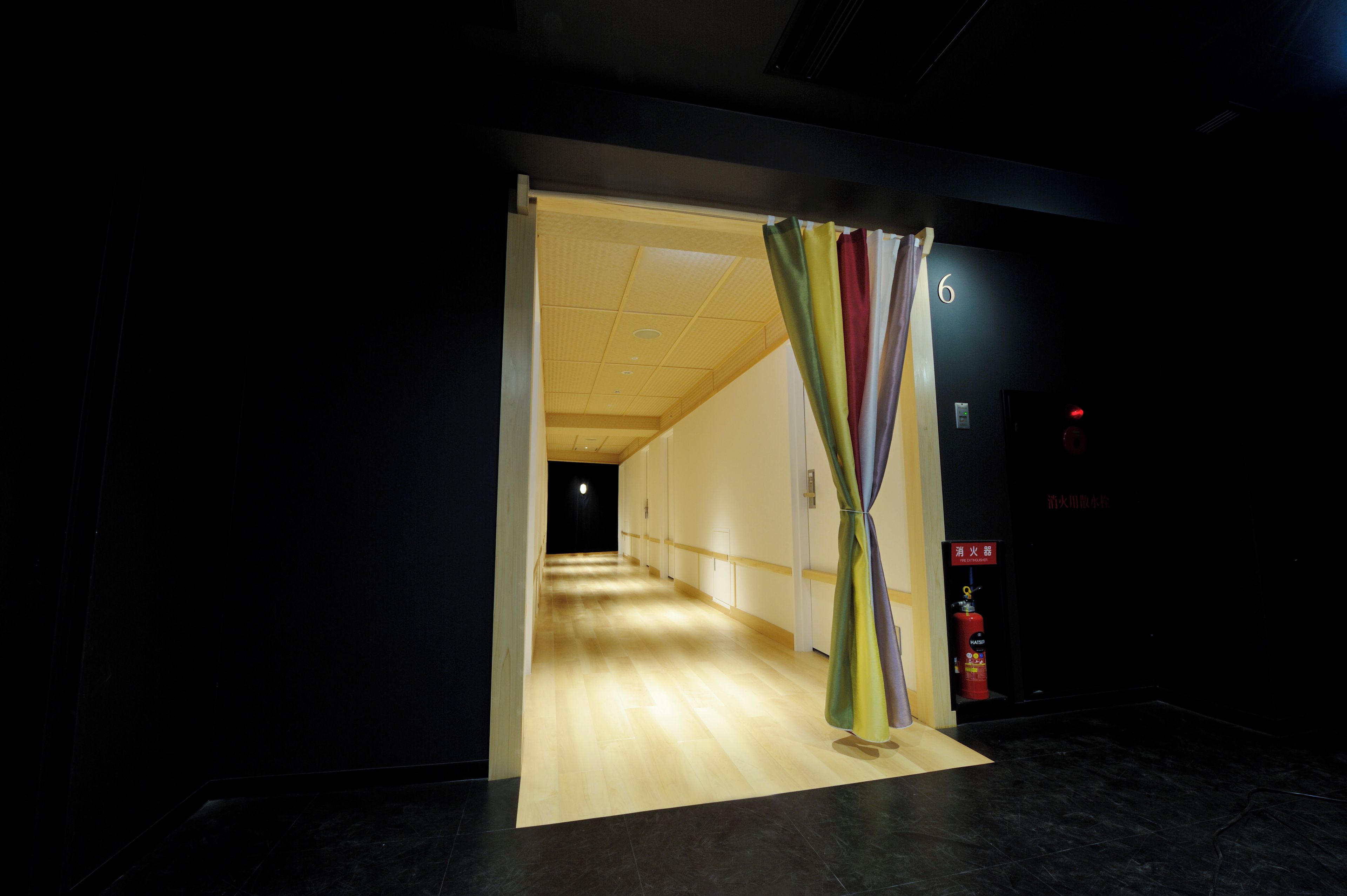 A corridor with a wooden floor leading to a room with a door. A fire extinguisher is mounted on the wall.