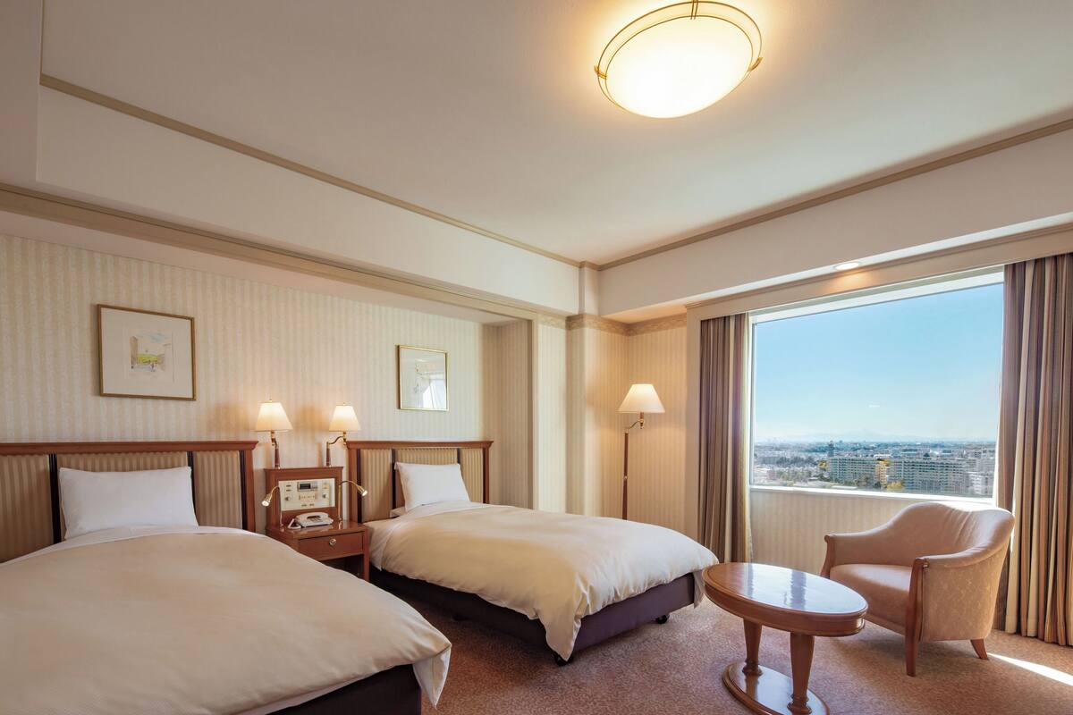 Hotelkamer met twee bedden, een tafeltje, een stoel, een nachtkastje met telefoon en een raam met uitzicht op de stad.