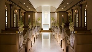 Interior de iglesia larga y elegante con bancas, arreglos florales y una cruz central.