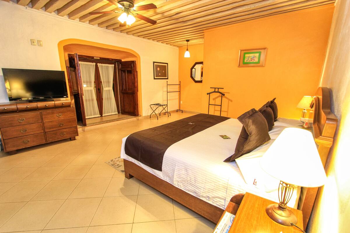 Una habitación de hotel con cama, televisión, cómoda de madera, lámpara y puerta que da a un balcón.