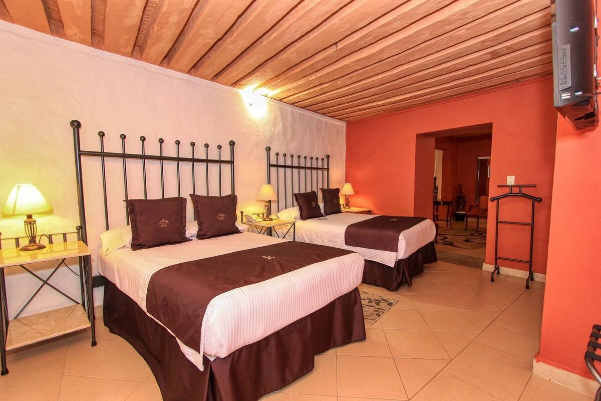 Habitación de hotel con dos camas, televisión, escritorio con lámpara y pared roja.
