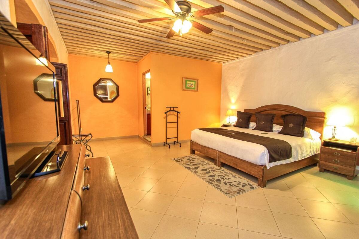 Un dormitorio con cama, cabecera de madera, mesitas de noche, ventilador de techo y televisor.