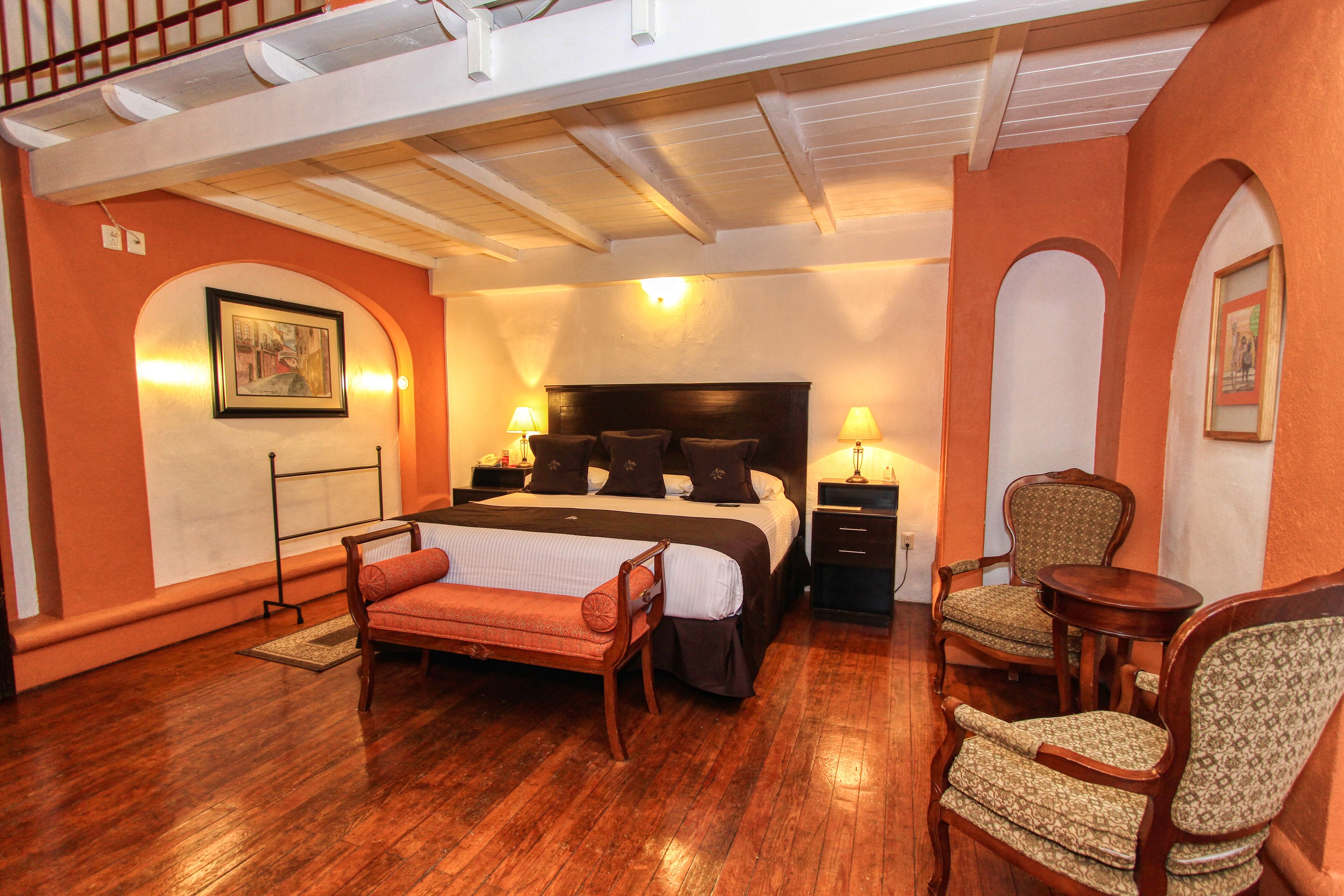Habitación de hotel con una cama grande, dos mesitas de noche con lámparas, un banco de madera, dos sillones tapizados y una pequeña mesa redonda.