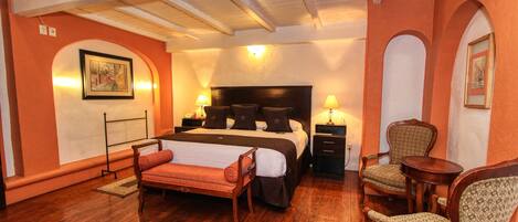 Habitación de hotel con una cama grande, dos mesitas de noche con lámparas, un banco de madera, dos sillones tapizados y una pequeña mesa redonda.