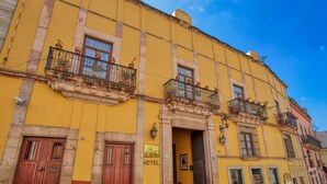 A yellow building with a sign indicating it is a hotel. There are multiple balconies with potted plants, and the entrance has a door and a window.