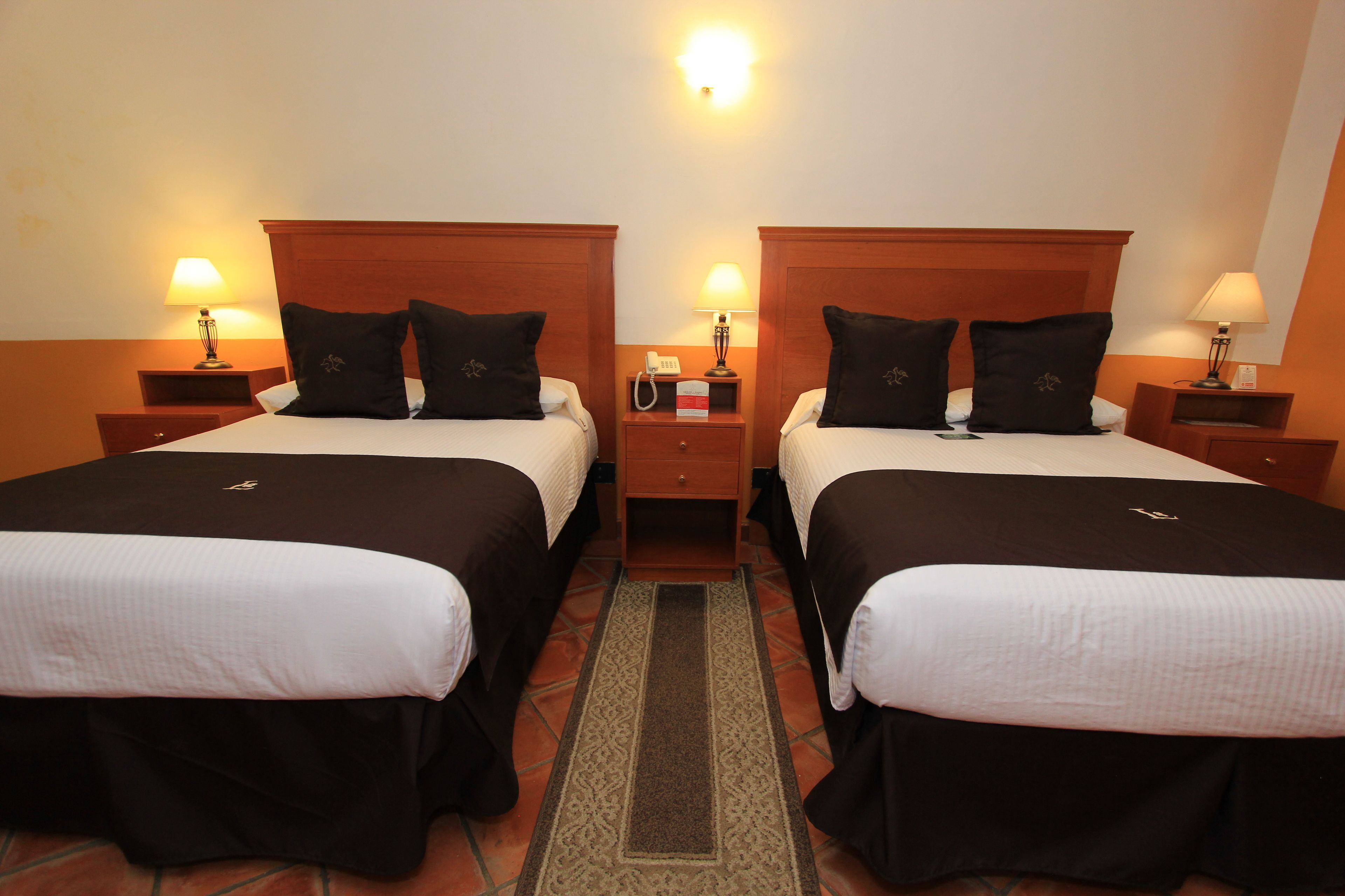 Habitación de hotel con dos camas, cada una con cabecero de madera, ropa de cama blanca y almohadas negras. Se observan mesitas de noche con lámparas, un buró con teléfono y un cajón pequeño.