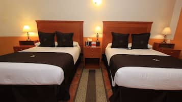 Habitación de hotel con dos camas, cada una con cabecero de madera, ropa de cama blanca y almohadas negras. Se observan mesitas de noche con lámparas, un buró con teléfono y un cajón pequeño.