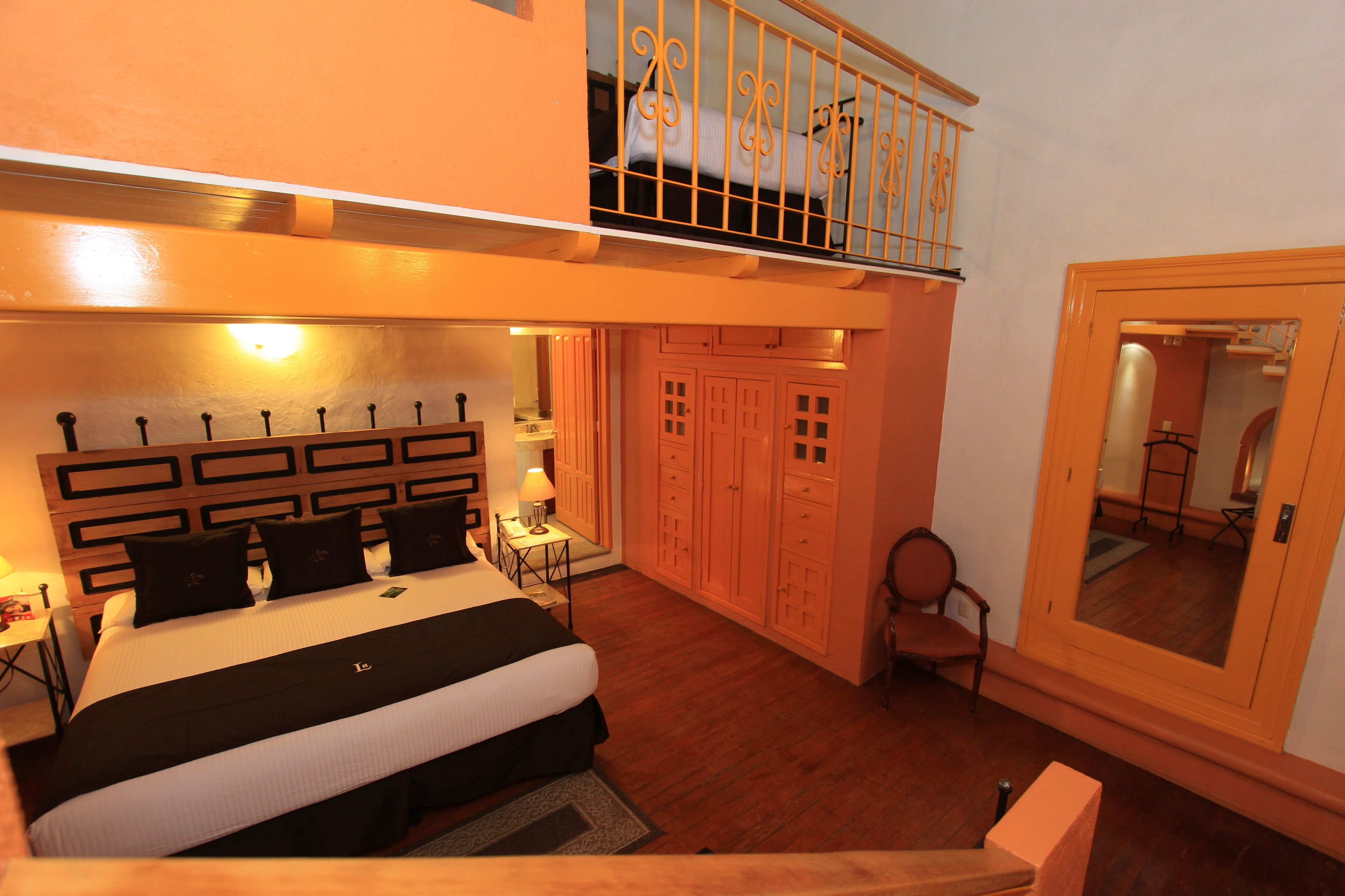 A bedroom with a large bed, a desk, a chair, and a small table. There is a balcony with a railing and a window.