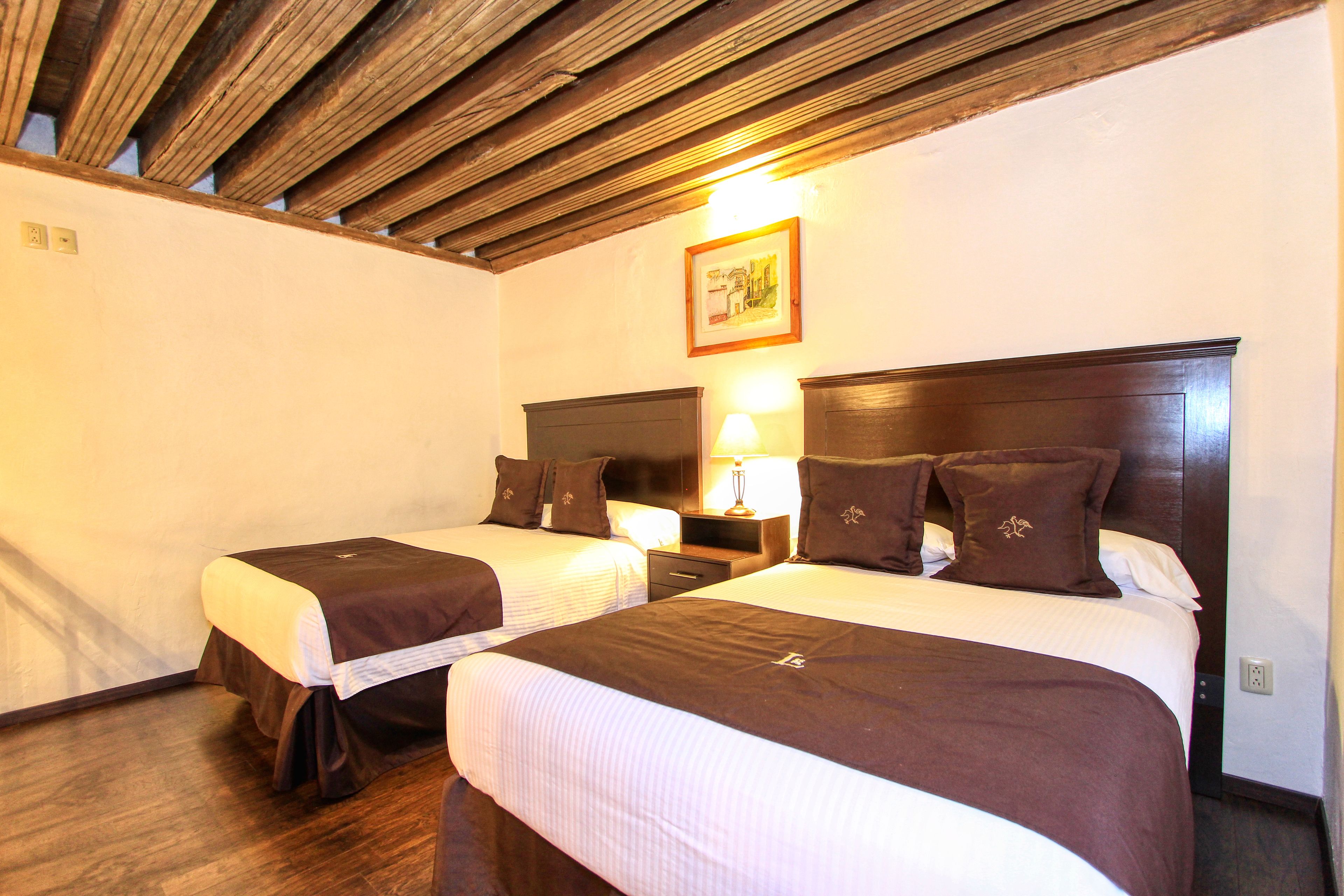 A hotel room with two beds, wooden ceiling beams, a nightstand with a lamp, and a framed picture on the wall.