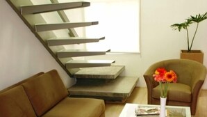 Uma sala de estar moderna com um sofá marrom, uma mesa de centro de vidro e uma poltrona. Uma escadaria é visível ao fundo.