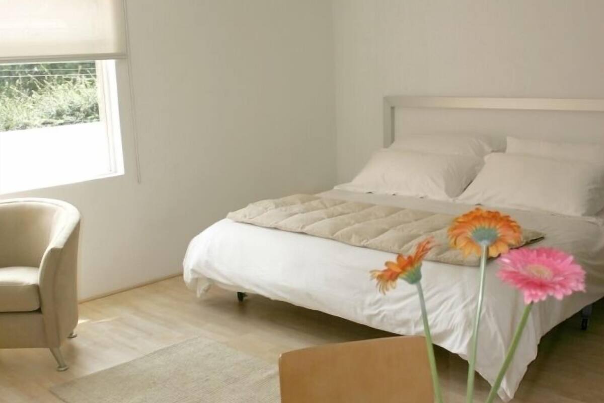 Ein Schlafzimmer mit Bett, einem Sessel, einem runden Tisch mit Blumenvase und einem Fenster mit Jalousie.
