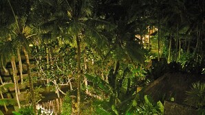 Vista noturna de um resort tropical com caminhos iluminados, palmeiras e uma ponte de pedra.