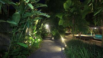 A nighttime pathway flanked by lush tropical plants, leading to a pool area with a covered seating area.
