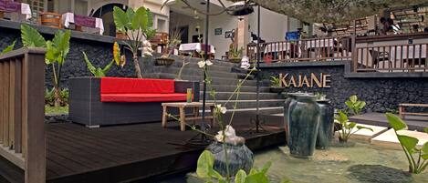 Um jardim interno tranquilo com um deck de madeira, um sofá vermelho e um elemento decorativo com água chamado "KAJANE".