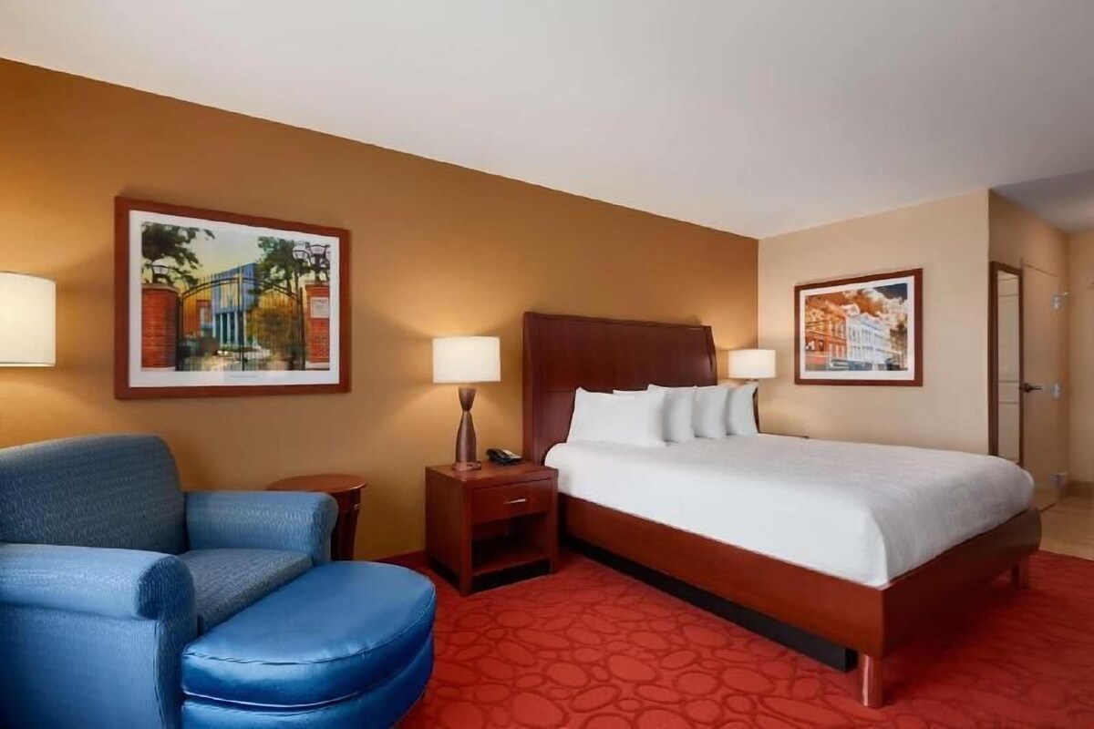 A hotel room with a bed, a blue armchair, a nightstand with a lamp, and two framed pictures on the walls.