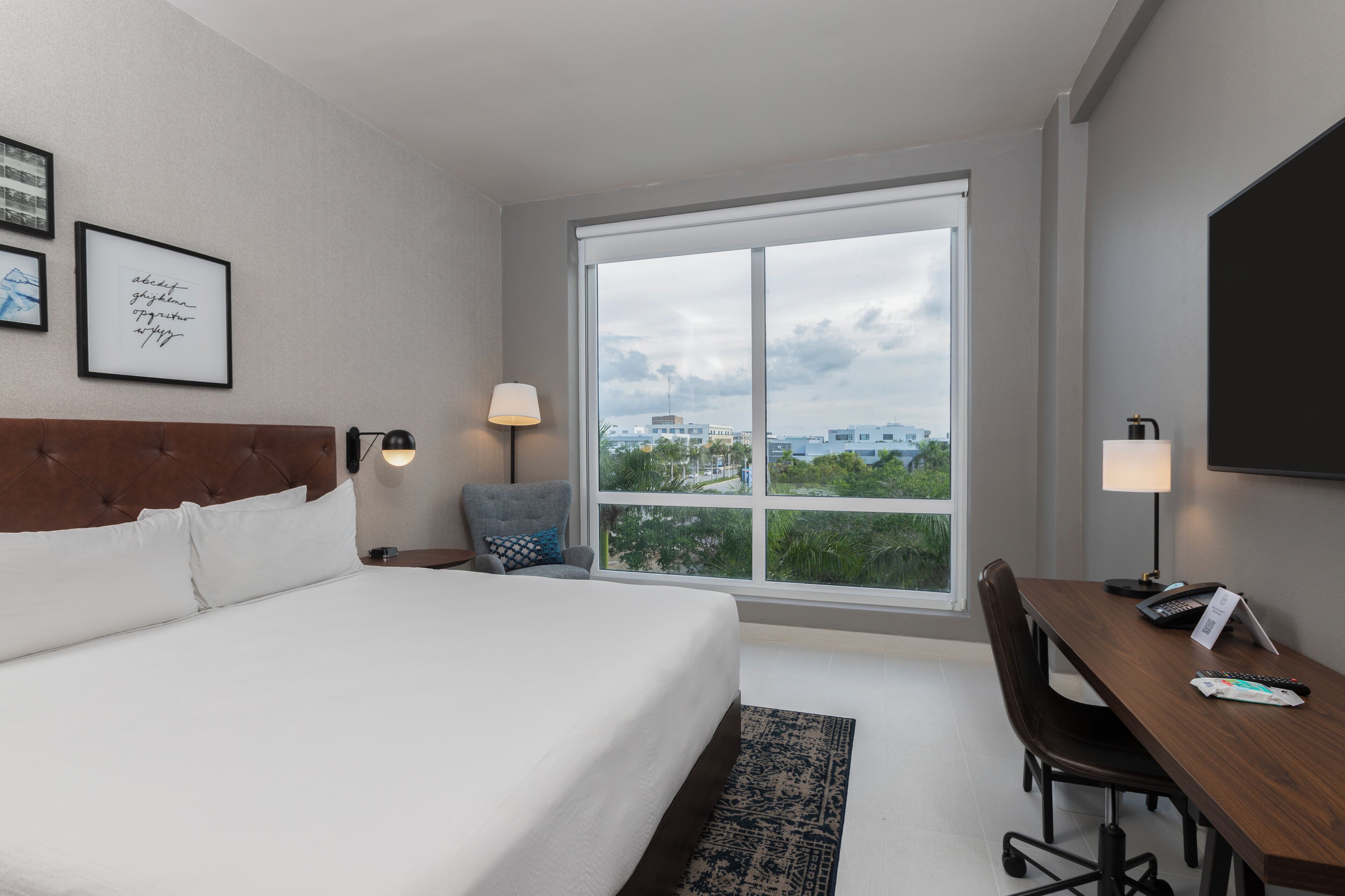 A hotel room with a large bed, a desk, a chair, a TV, and a window with a city view.