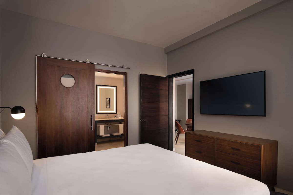 Une chambre d’hôtel avec un lit, une commode, une télévision, un miroir et une salle de bain visible à travers une porte ouverte.