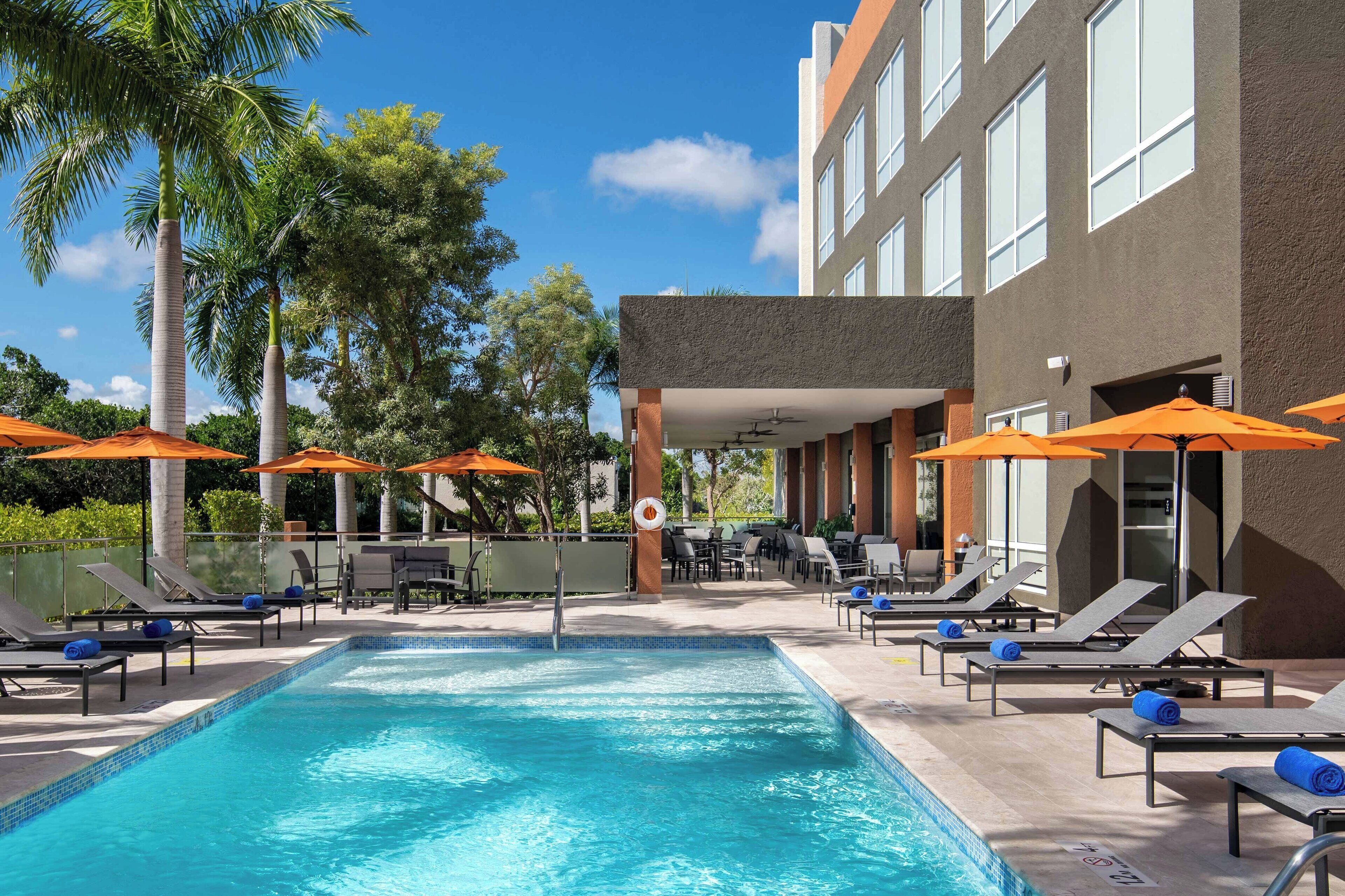 Una piscina con sillones de descanso y sombrillas, rodeada de palmeras y un edificio con amplias ventanas.