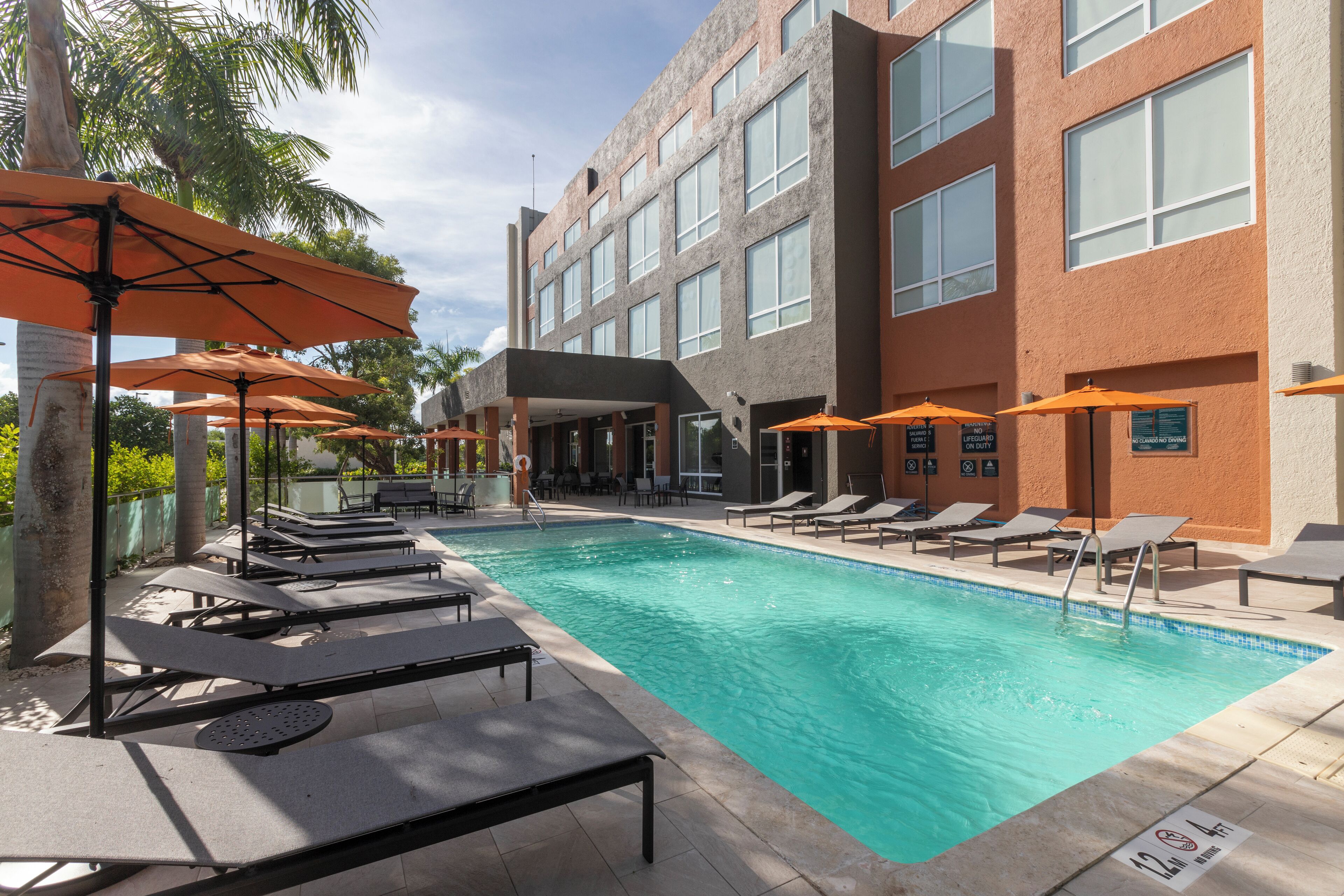 Una piscina con tumbonas y sombrillas, rodeada por un edificio con múltiples ventanas y una zona cubierta.