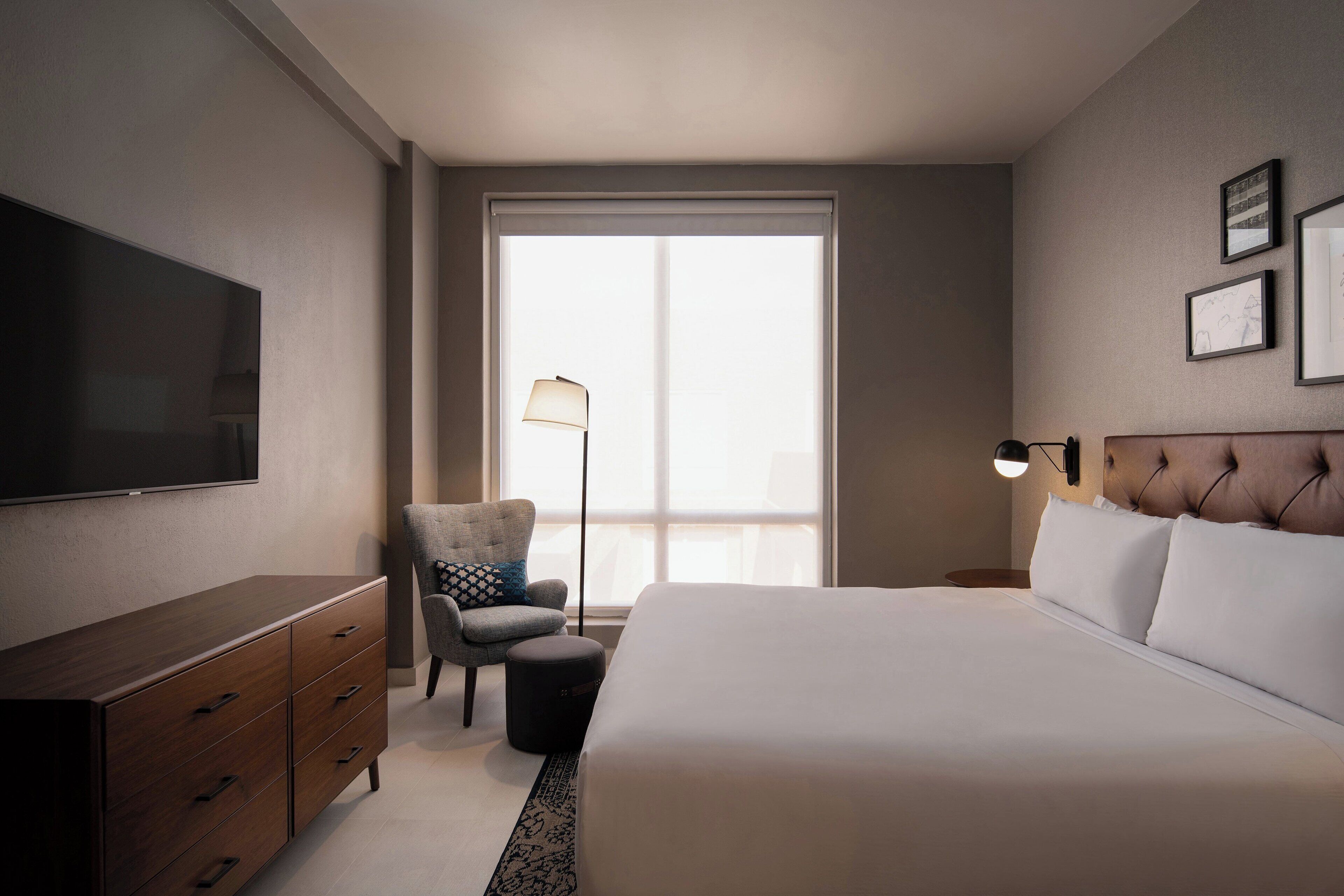 Habitación de hotel con una cama grande, televisor de pantalla plana, una silla, un escritorio y una ventana con persianas.