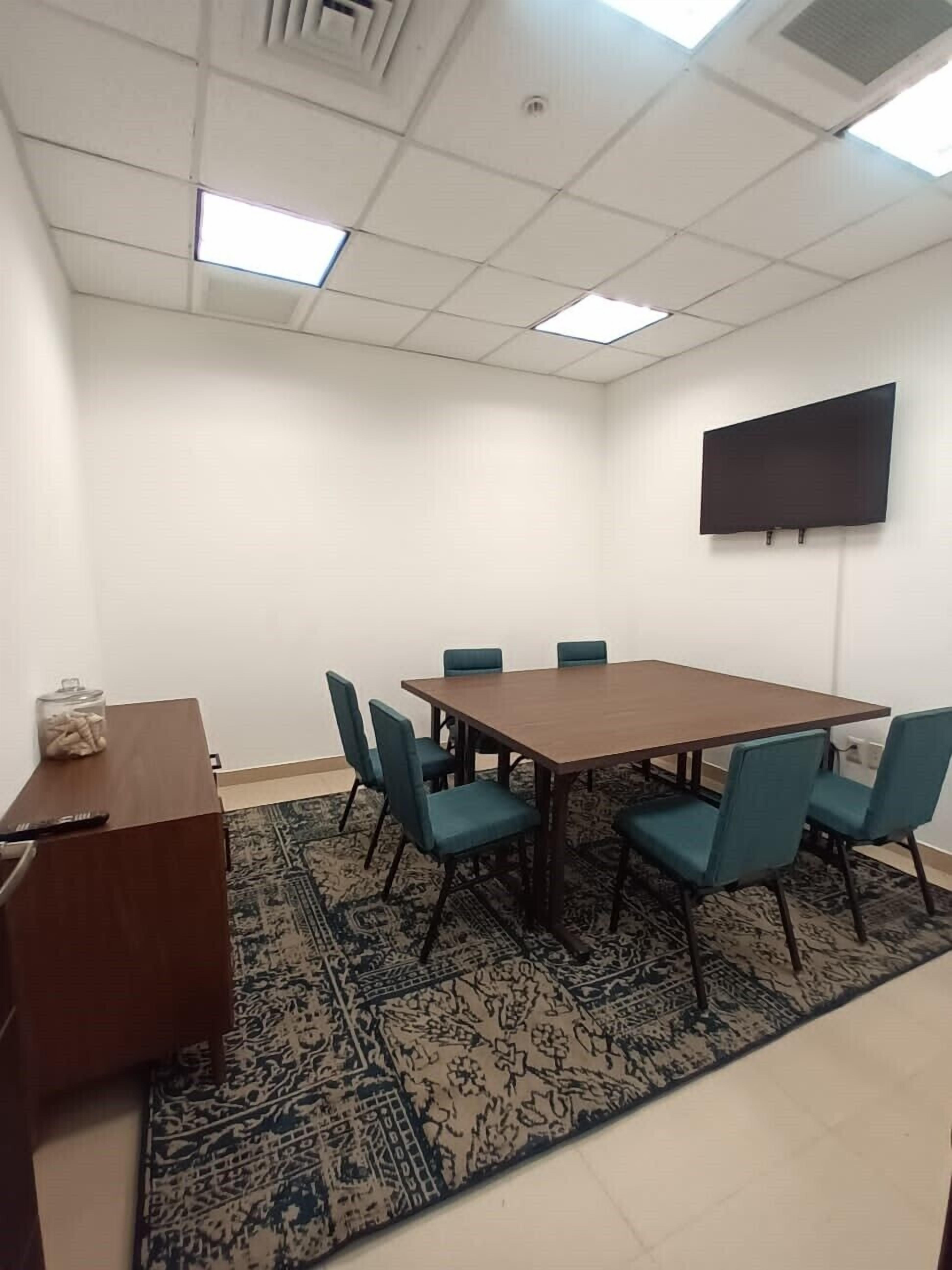 A conference room with a large table, six chairs, a small desk with a computer, and a TV on the wall.