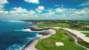 Un parcours de golf situé sur la côte, offrant une vue dégagée sur l’océan, une plage de sable et un terrain verdoyant parfaitement entretenu.