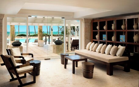 A spacious living area with a large sofa, armchairs, and a coffee table. A glass door leads to an outdoor pool area with lounge chairs and a view of the ocean.