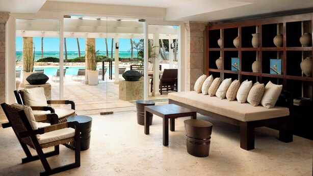 Un vaste espace de vie comprenant un grand canapé, des fauteuils et une table basse. Une porte vitrée donne accès à un espace extérieur avec une piscine, des chaises longues et une vue sur l’océan.