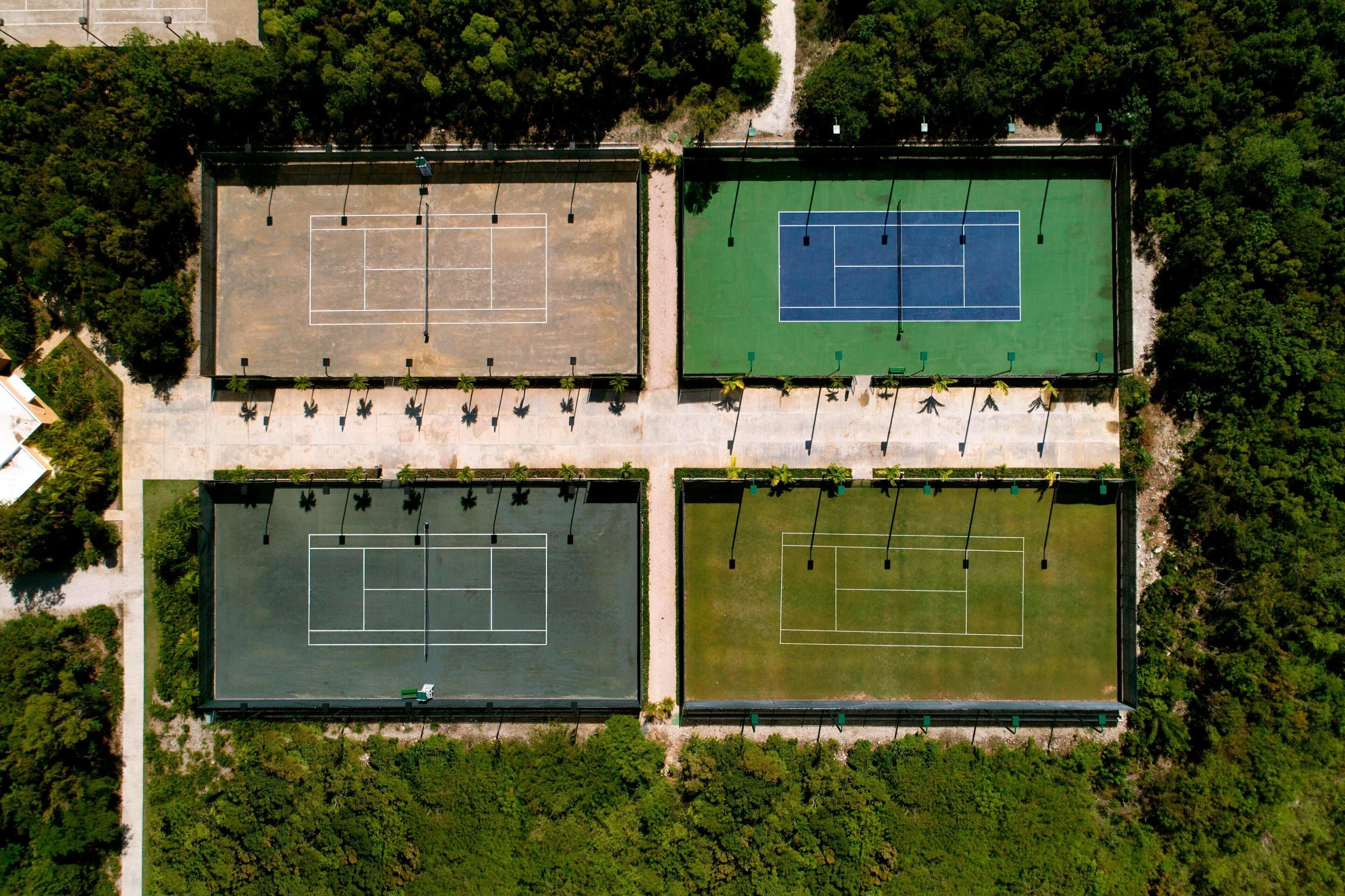 An aerial view of a tennis court complex with four courts, surrounded by dense greenery.