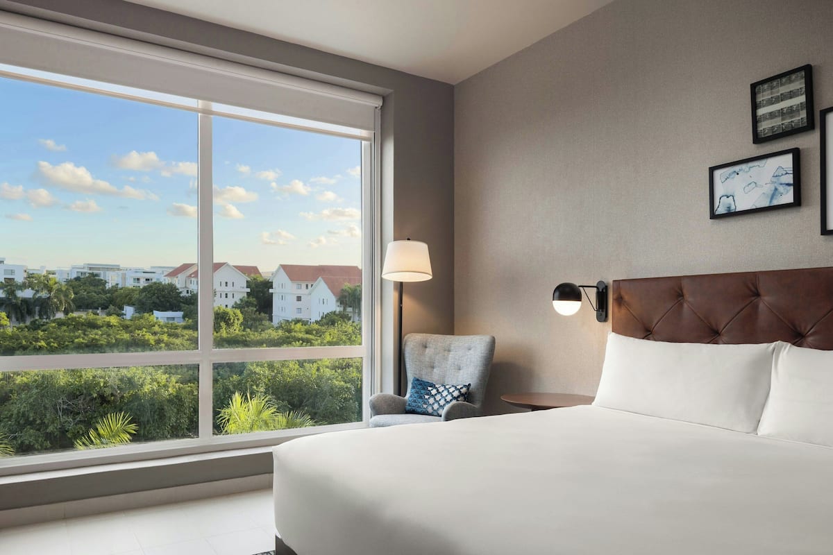 Une chambre d’hôtel dotée d’une grande fenêtre, d’un lit avec du linge de maison blanc, d’une tête de lit capitonnée, d’une table de chevet avec une lampe et d’une chaise agrémentée d’un coussin à motifs.