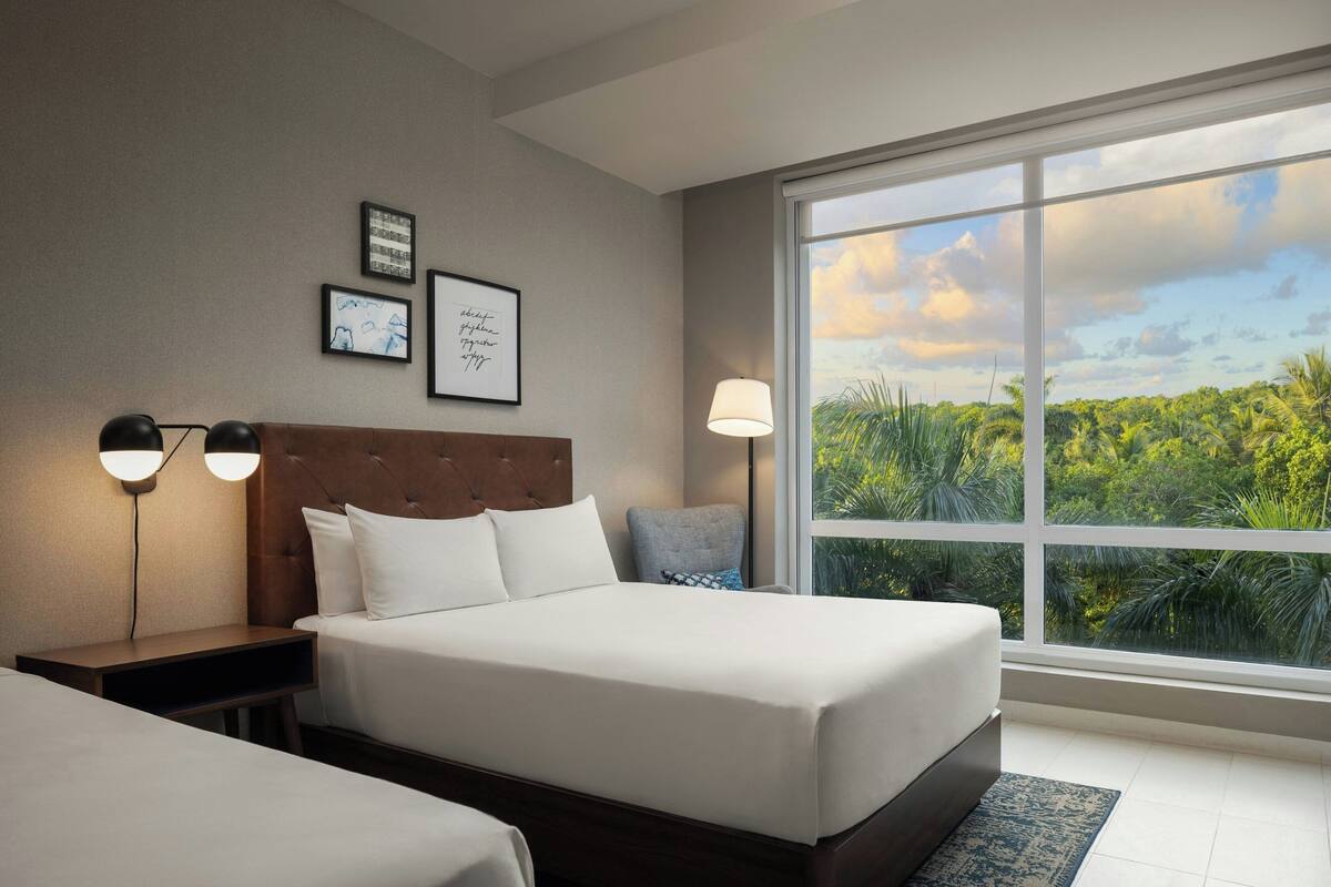 Une chambre d’hôtel avec une grande fenêtre, un lit recouvert de draps blancs, une table de chevet avec une lampe et des œuvres d’art encadrées accrochées au mur.