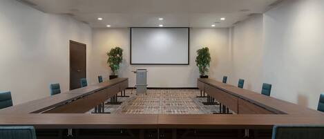 A conference room with a long table, chairs, a projector screen, and potted plants.