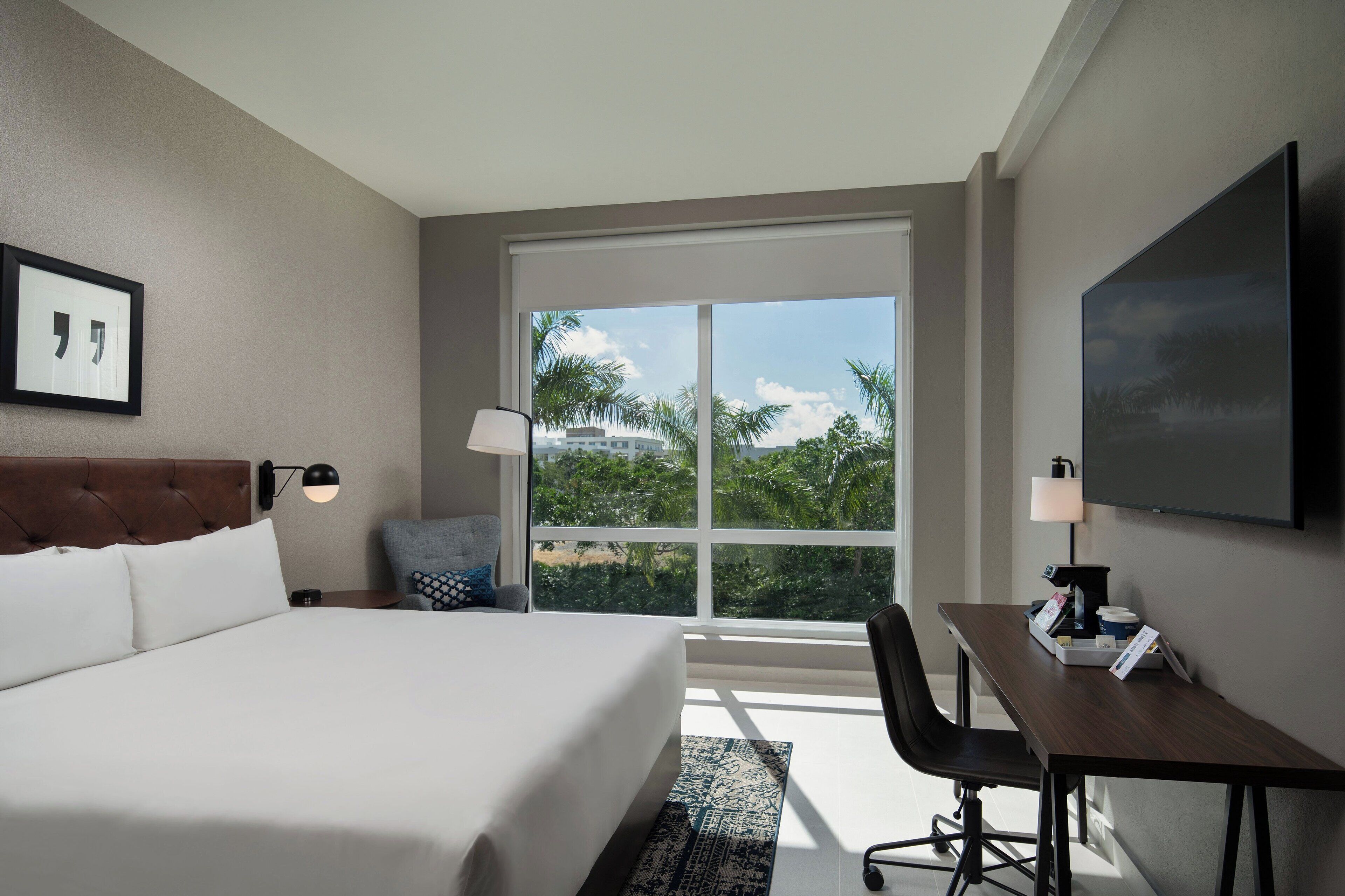 A hotel room with a large bed, a desk, a TV, and a window with a view of palm trees and buildings.
