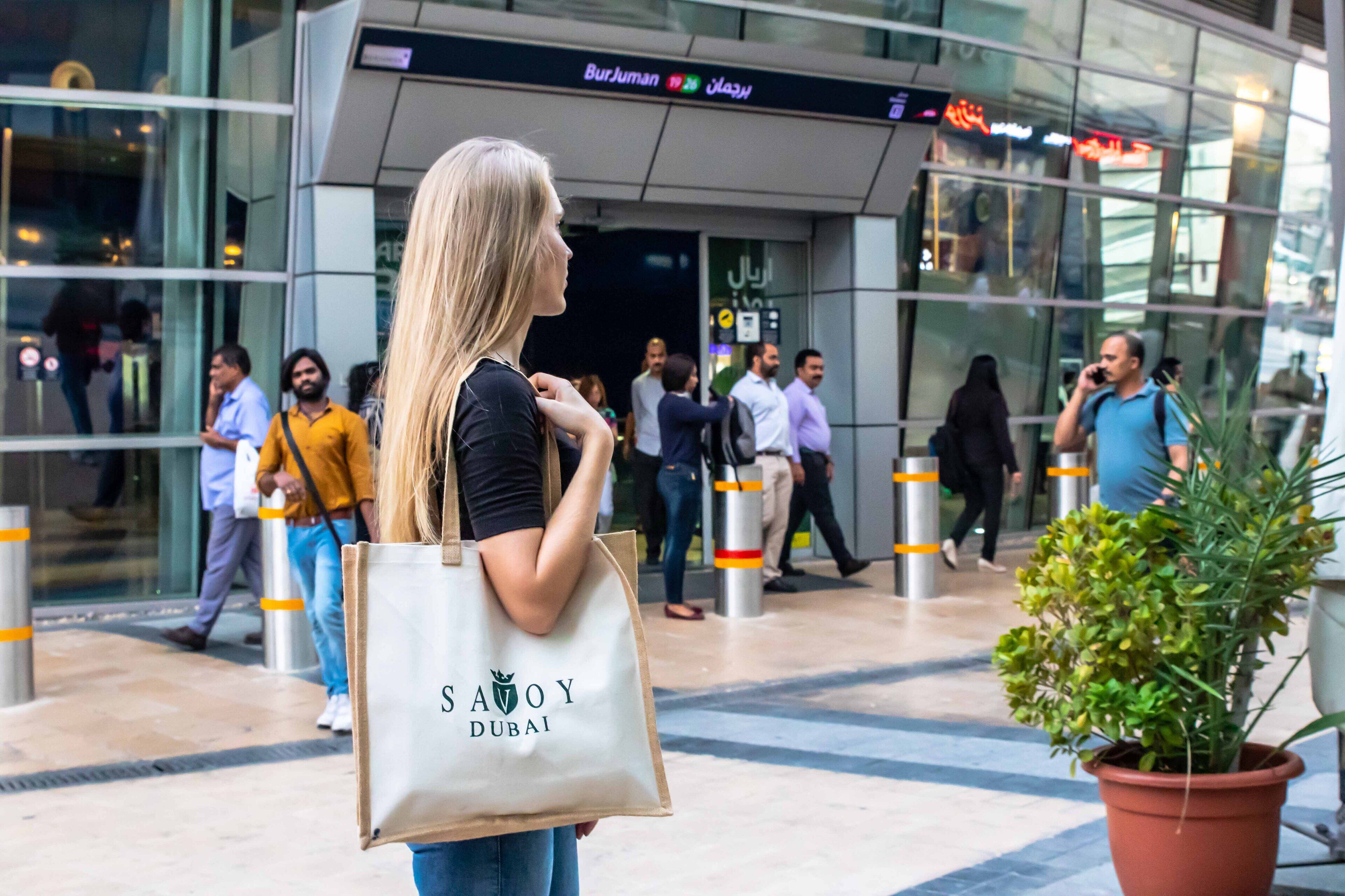 A woman holding a 'SAYOY DUBAI' tote bag in front of a modern building with glass windows and a sign indicating 'Burjuman' and 'Dubai'.