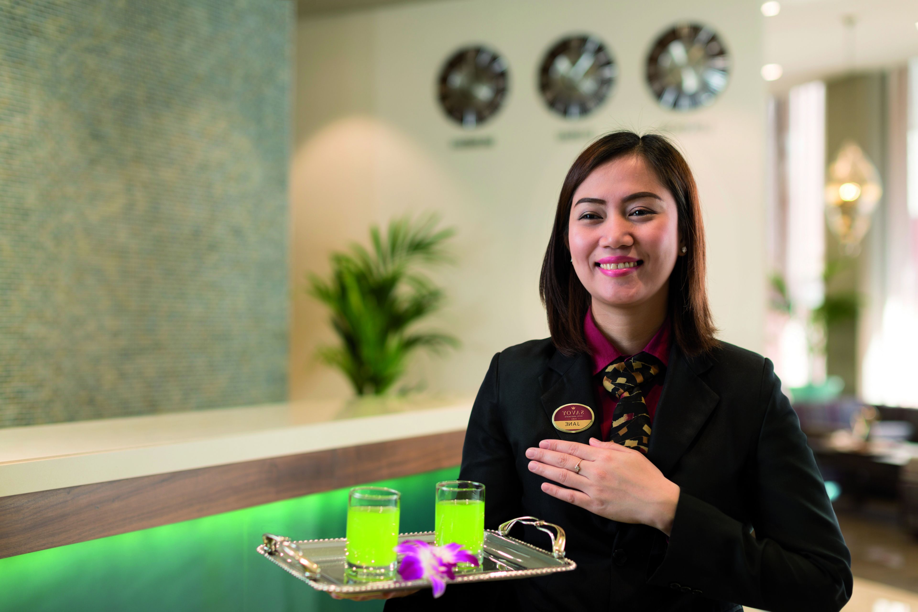 A person holding a tray with two glasses of green beverage and a small round object, in a room with a green accent wall and clocks on the wall.