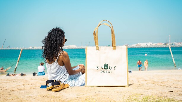 Une scène de plage avec une personne assise sur le sable, un sac de plage portant l’inscription « SAVOY DUBAI », et l’océan en arrière-plan.
