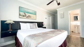 A hotel room with a bed, a nightstand, a lamp, a ceiling fan, a painting of a beach scene, a door, and a bathroom visible through an open door.