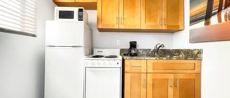 A compact kitchen with white appliances, wooden cabinets, and a painting above the sink.