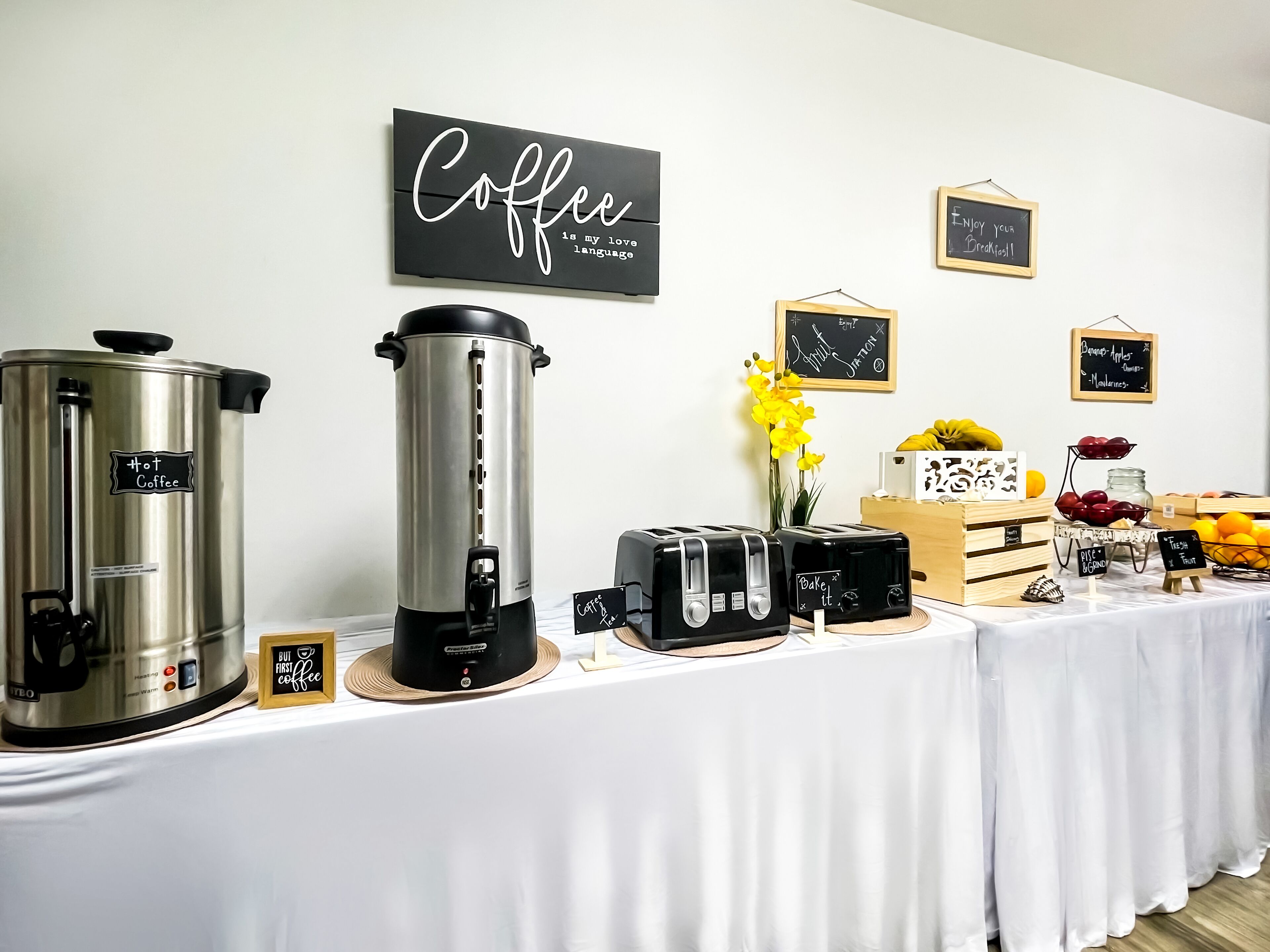 A coffee station with a large coffee pot, a coffee maker, and a toaster. There are also signs with coffee-related messages and a display of fruits and vegetables.