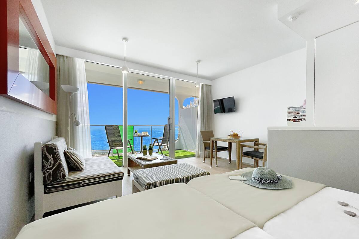 A hotel room with a large bed, a small table with chairs, a TV on the wall, and a balcony with a view of the sea.