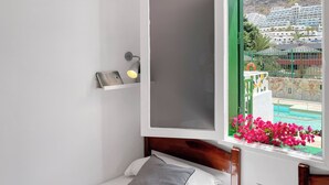 Een slaapkamer met een bed, nachtkastje met een lamp, raam met uitzicht op een zwembad en gebouwen.