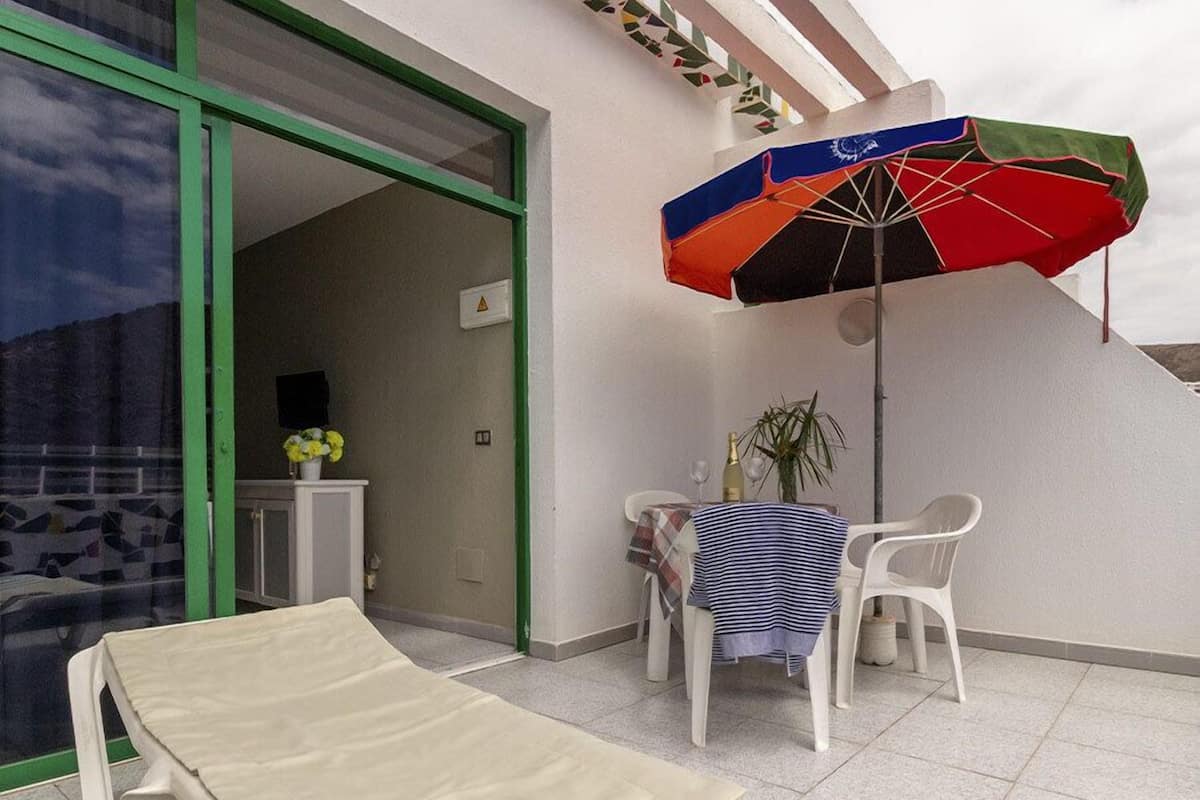 A small outdoor seating area with a table and chairs, a colorful umbrella, and a view of the interior through a glass door.
